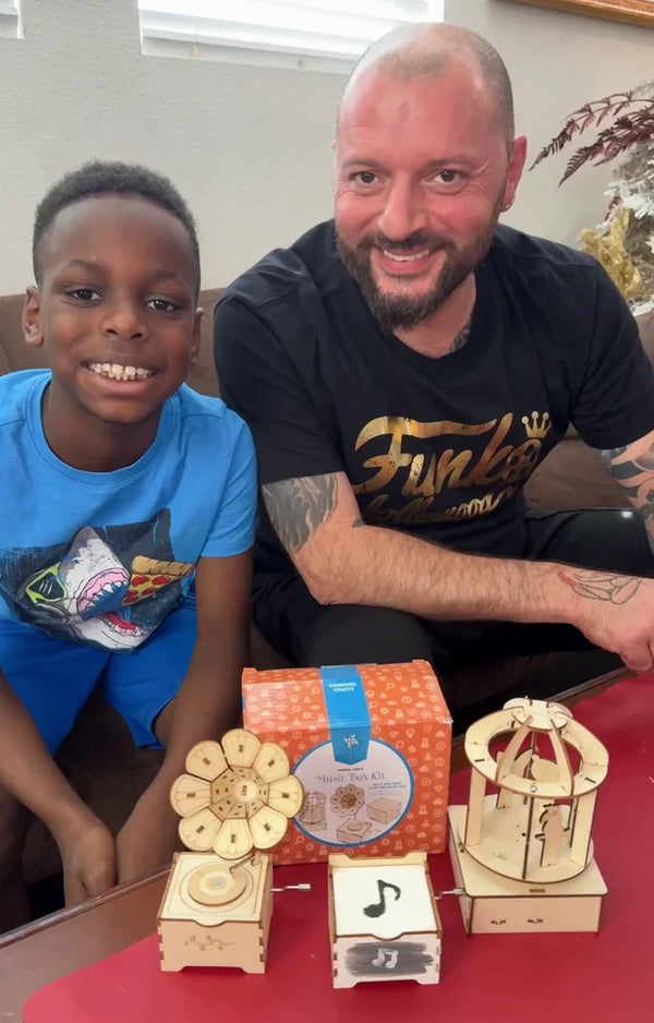 Smiling boy and his dad sitting behind the three wooden music boxes they built and the kit box