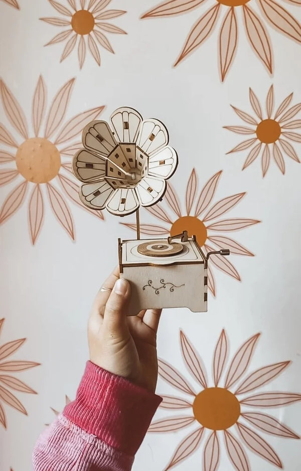 A child's hand holding up a finished wooden flower gramophone music box against a daisy wallpaper