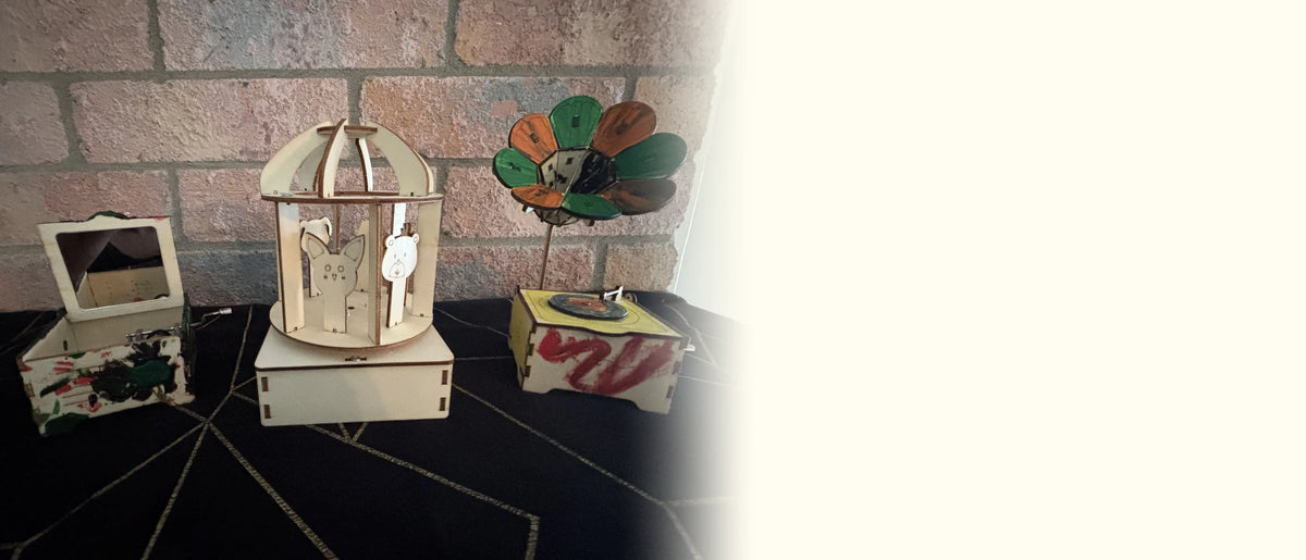 Three painted wooden music boxes, a mirror box, a carousel and a flower gramophone, against a brick wall