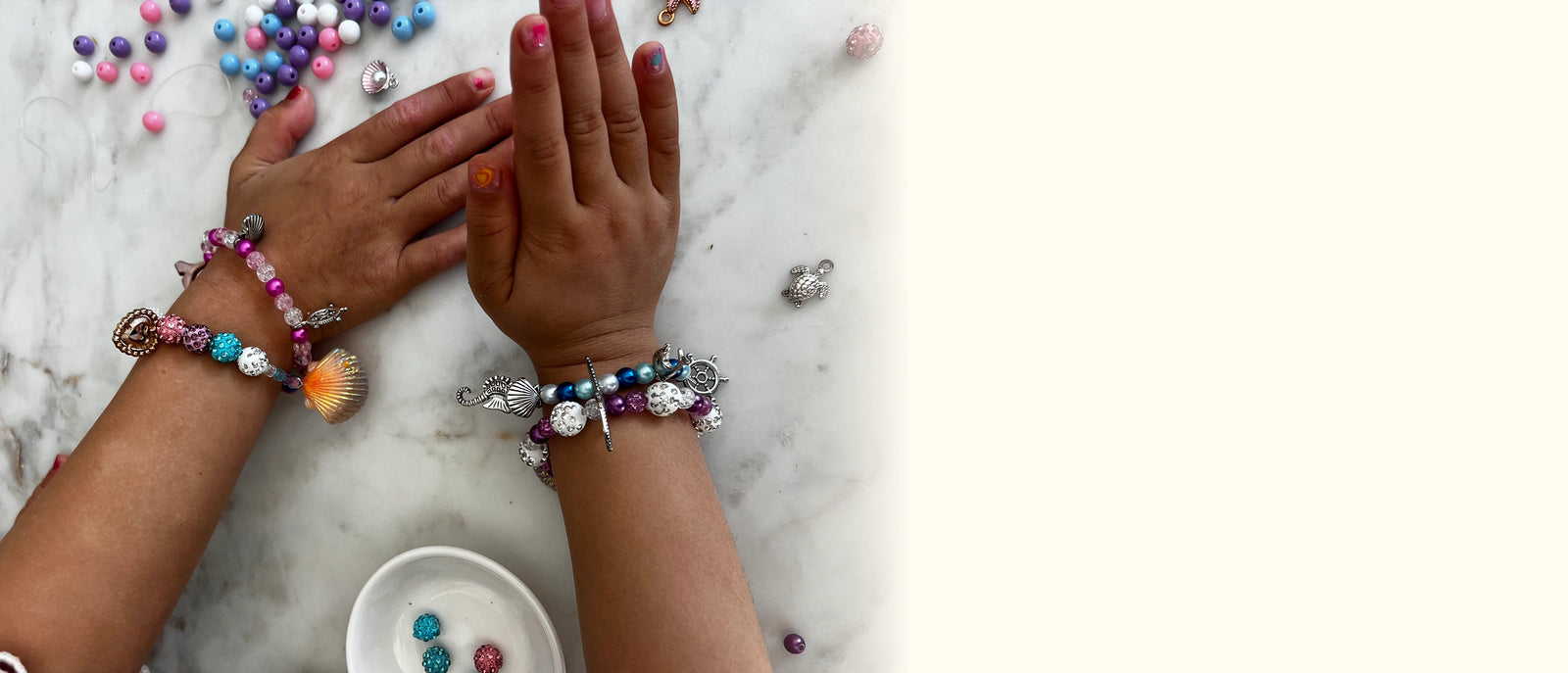 A top-down view of two hands wearing colorful beaded bracelets on a white marble surface, with loose beads and a small dish nearby.