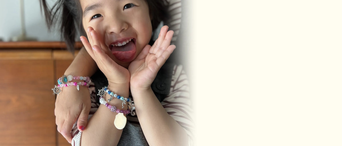 Grinning girl with her hands on her cheeks, wearing three mermaid charm bracelets she made