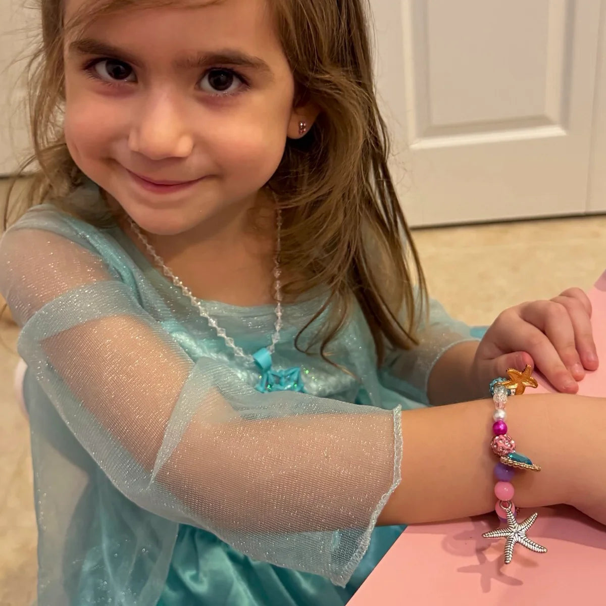Girl in a blue dress smiling at a pink desk, wearing the mermaid charm bracelet she made