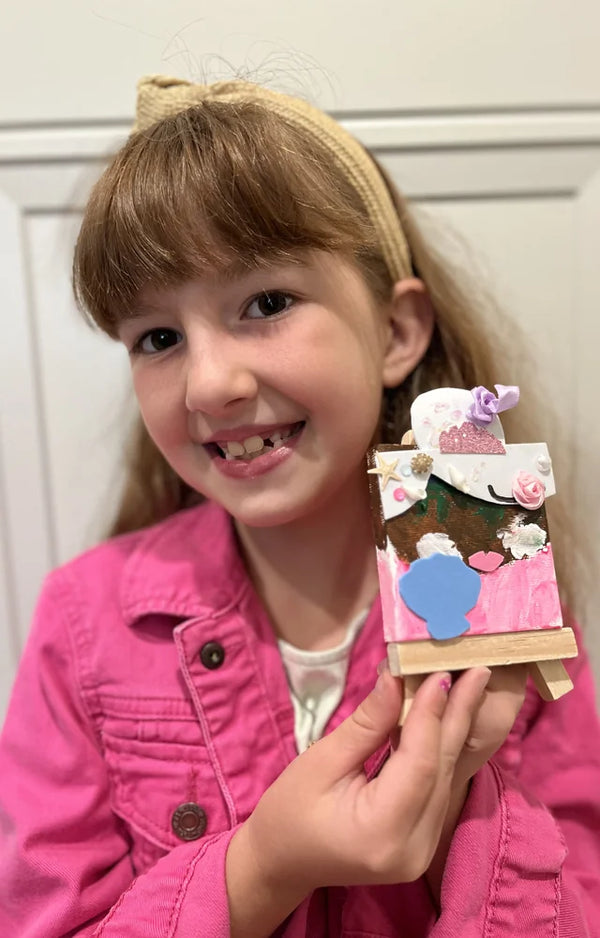 A girl in a pink jacket holds her unicorn canvas decorated with fabric flowers and a seashell
