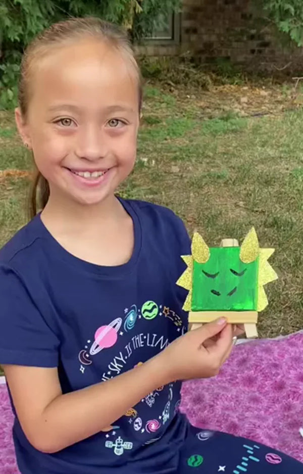 A girl outdoors shows her finished green dragon canvas standing on its little wooden easel