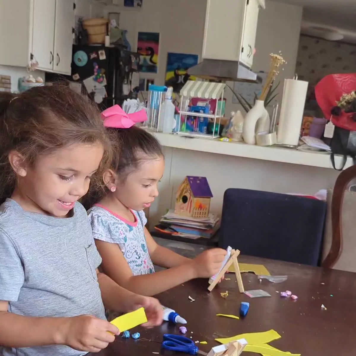 Two sisters smile while they cut and glue decorations onto their mini canvases at the kitchen table