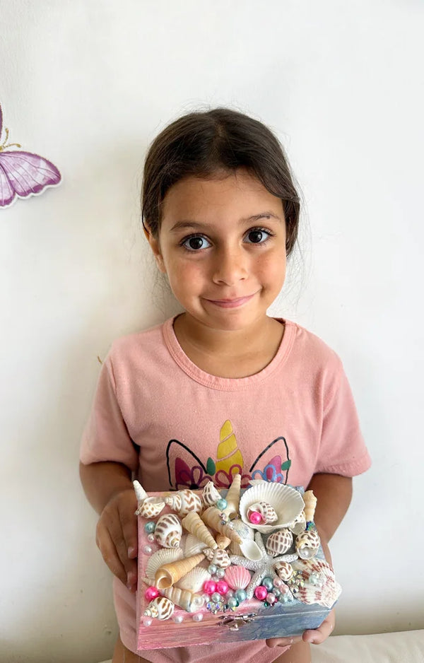 Girl in a unicorn tee holding her finished jewelry box covered in sea shells