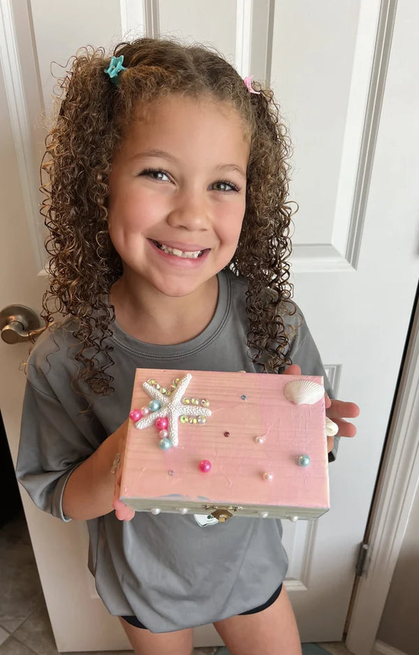 Girl with curly hair grinning, holding her pink jewelry box decorated with a starfish