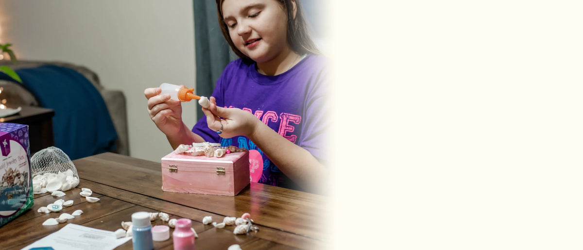 Girl gluing a sea shell beside her pink mermaid jewelry box, with the kit box on the table