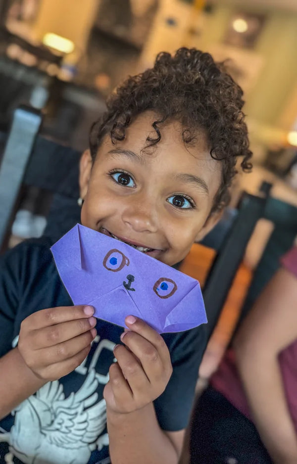 A smiling boy with curly hair holding a purple folded paper face with drawn eyes in front of his own
