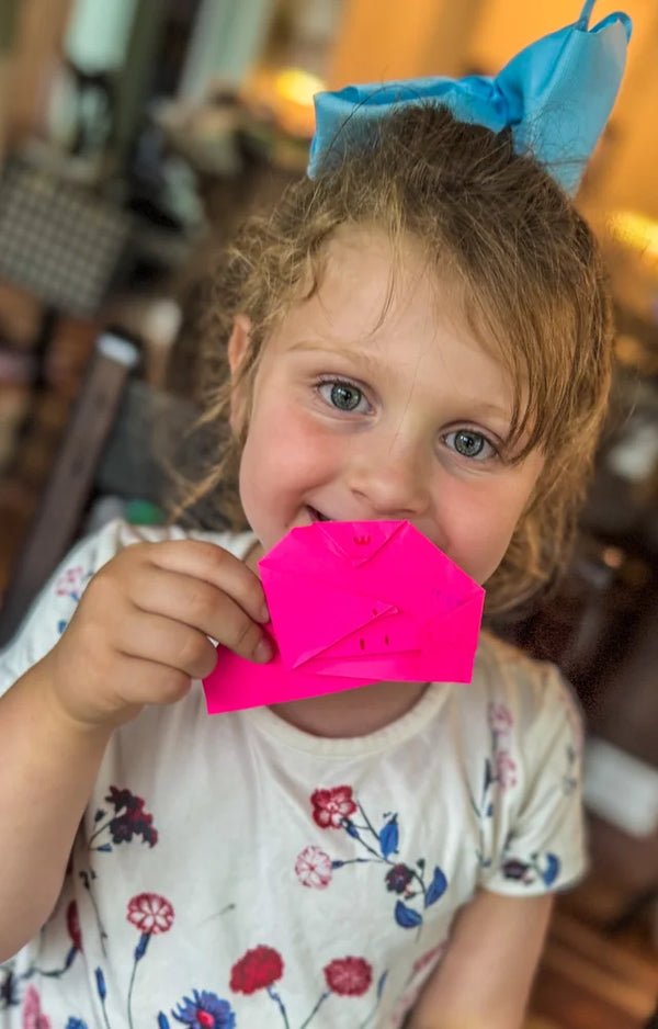 A girl with a blue hair bow holding a pink folded paper face up to her smile at the table