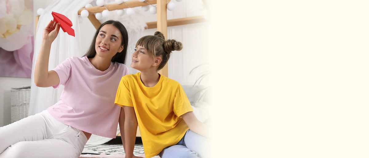 A woman and a girl in a yellow shirt sitting on the floor as the woman launches a red paper airplane