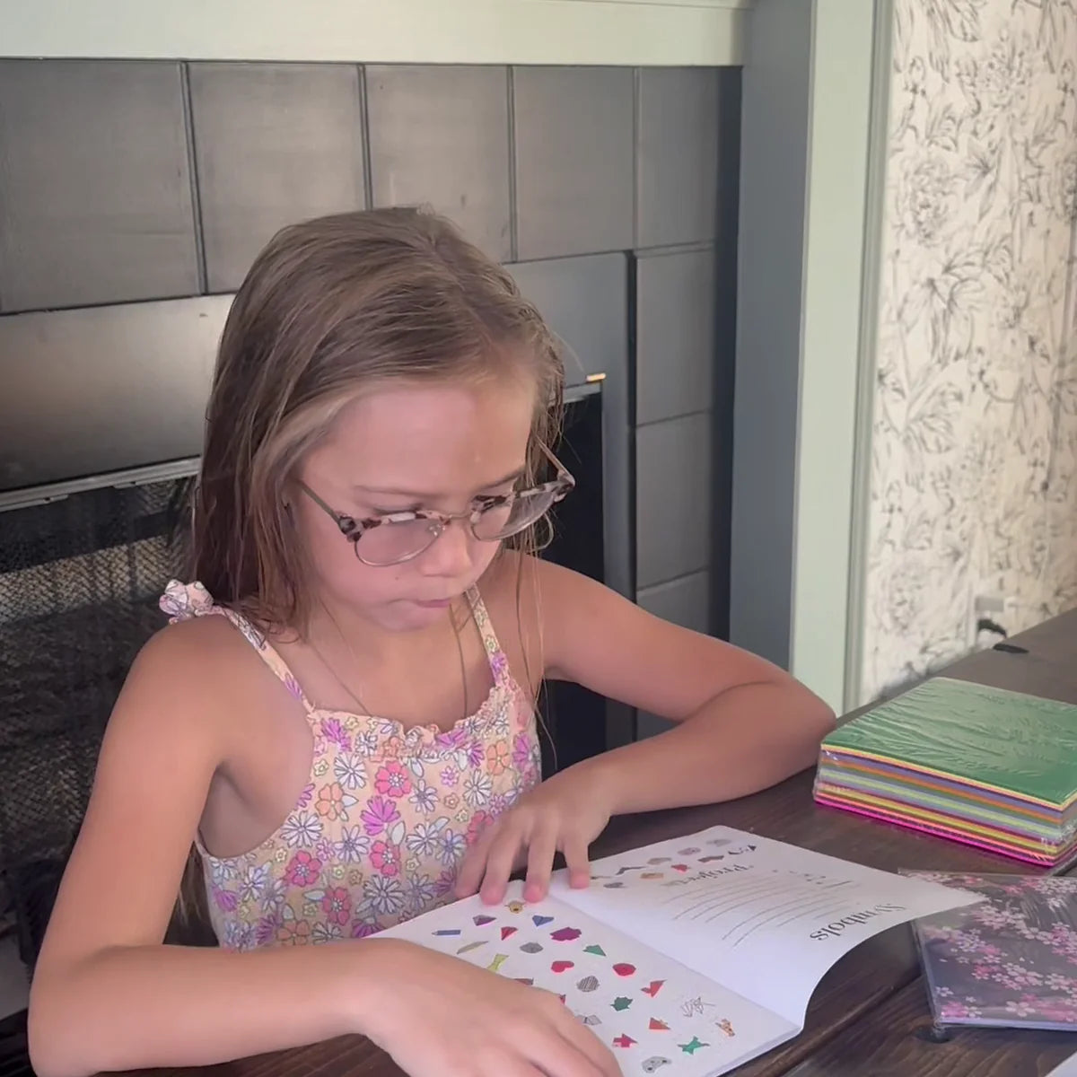 A girl in glasses reading the origami project book at a table with a stack of colored paper beside her