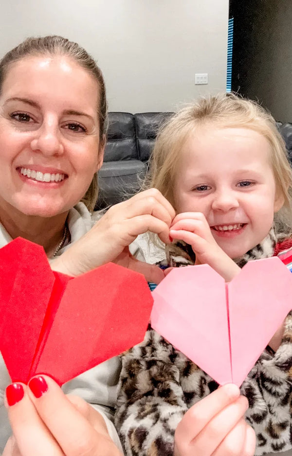 A smiling mother and young daughter holding up a red paper heart and a pink paper heart they folded