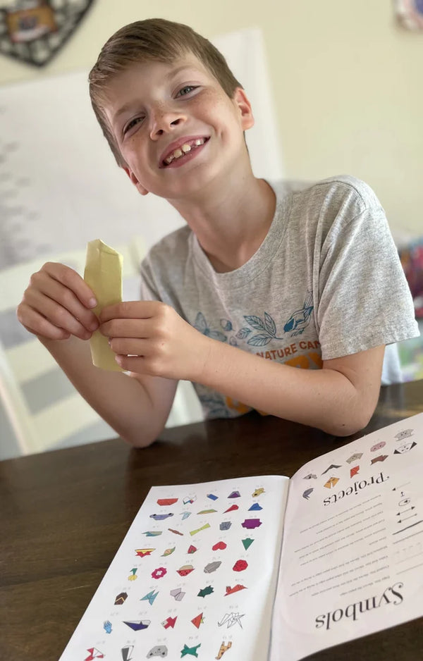 A grinning boy holding a folded yellow paper model above the open origami project book at a table