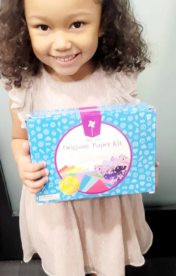 A smiling girl with curly hair in a pink dress holding the blue Origami Paper Kit box with both hands