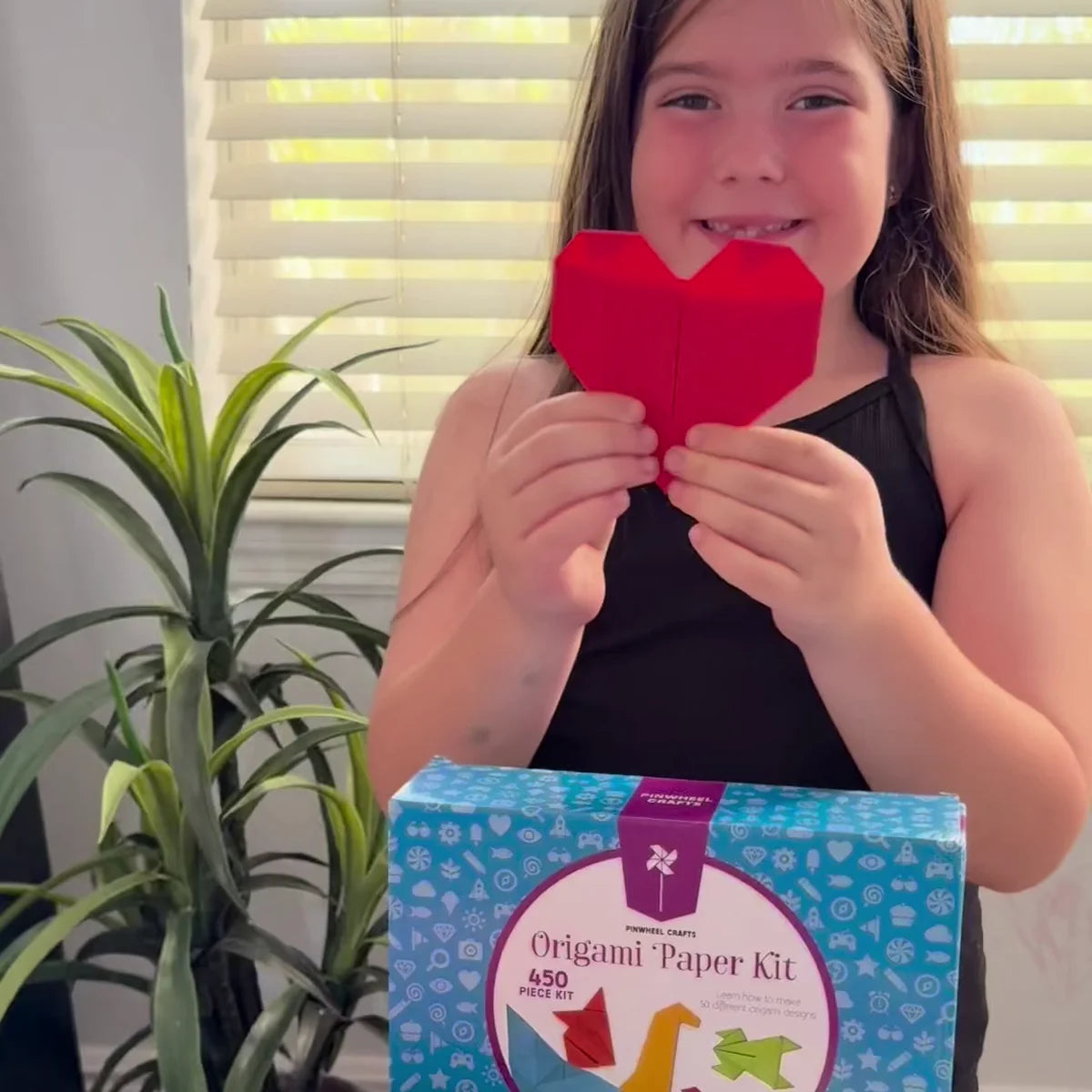 A smiling girl holding up a red folded paper heart behind the blue Origami Paper Kit box
