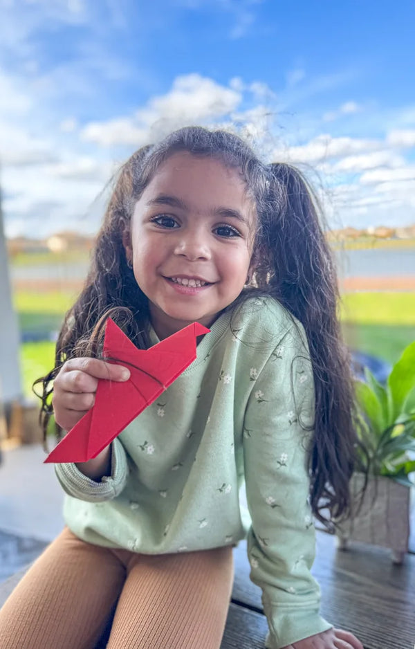 A smiling girl with long curly hair sitting outdoors and holding up a red folded paper model