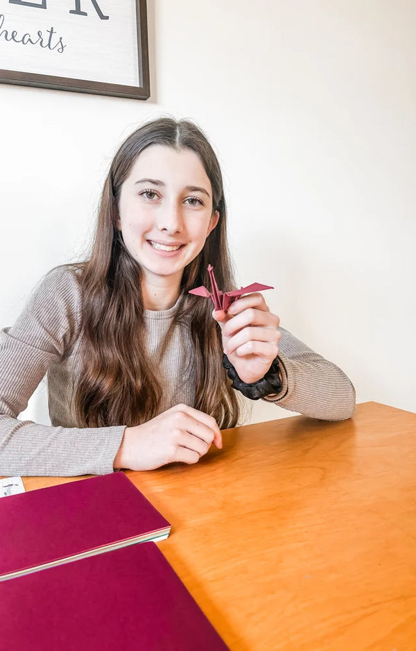 A smiling teen at a wooden table holding a small red paper crane beside sheets of folding paper