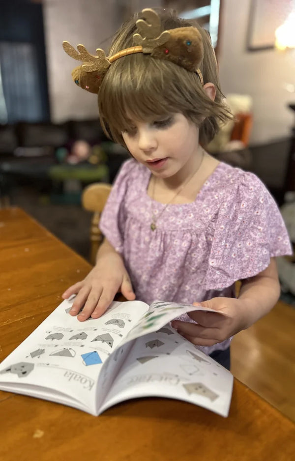 A young girl wearing a reindeer headband reading an origami project book at a wooden table