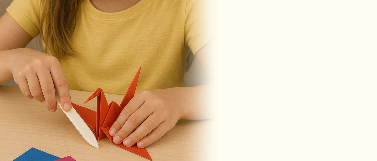 A child in a yellow shirt using a white bone folder to crease a red origami crane on a wooden table