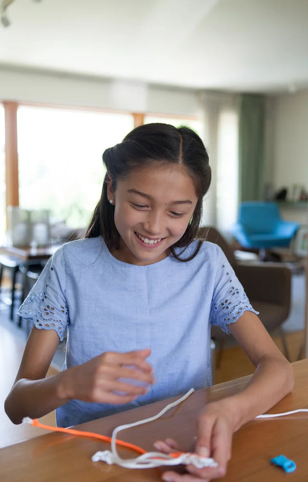 Girl smiling at the table while she weaves white and orange paracord into a bracelet