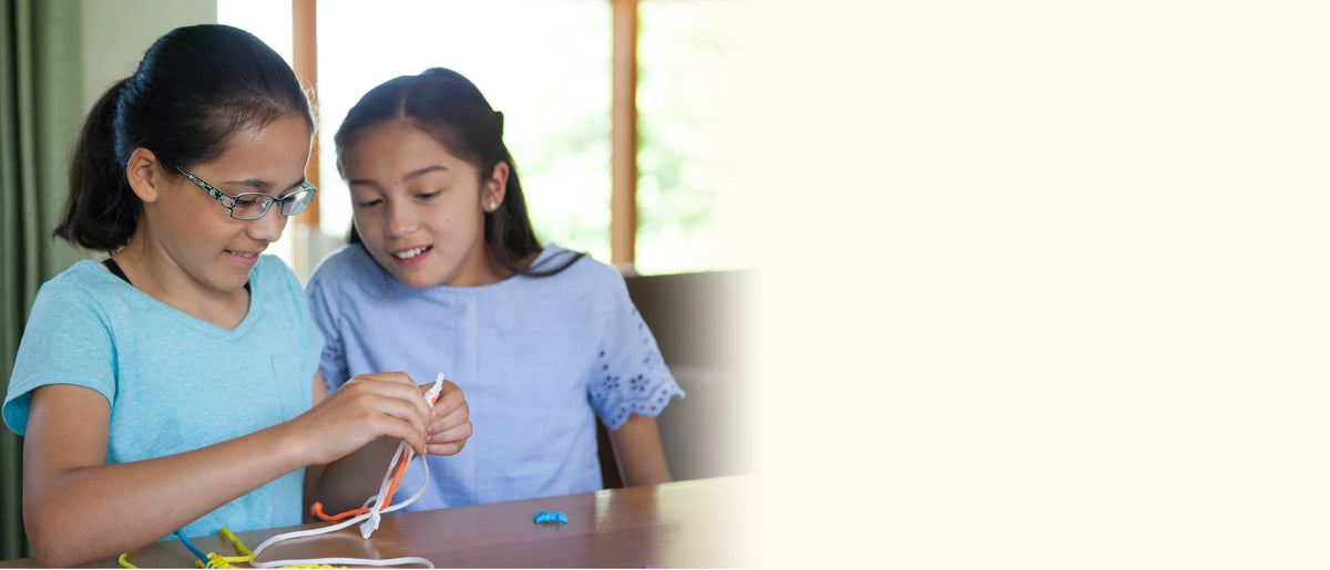 Two girls sitting at a wooden table weaving white and orange paracord into bracelets