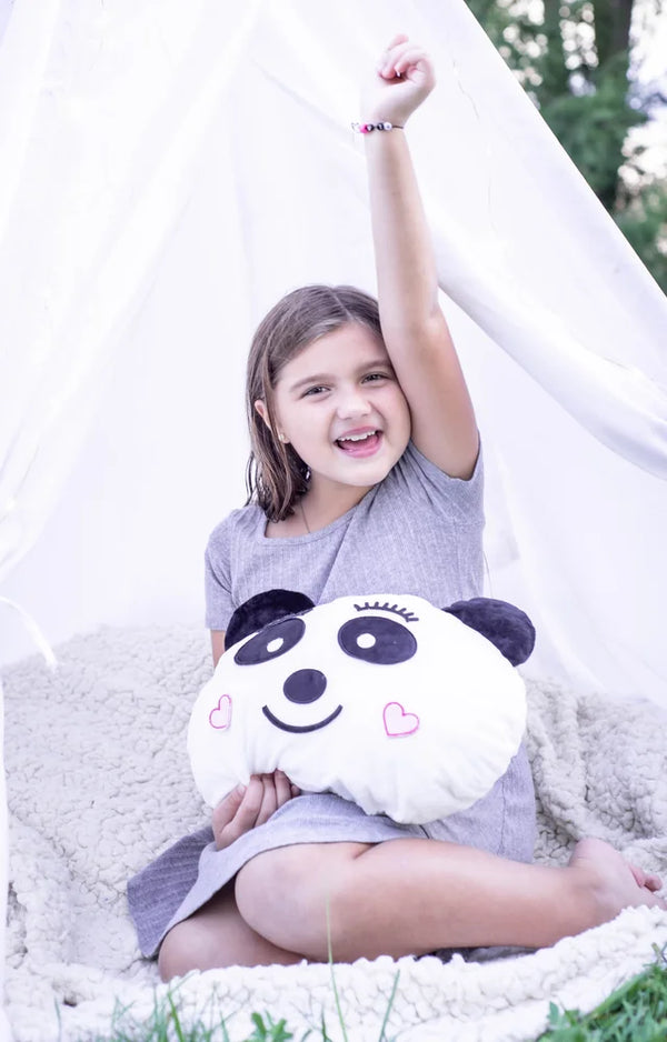 A girl in a white teepee raises her arm, with the finished panda pillow resting in her lap