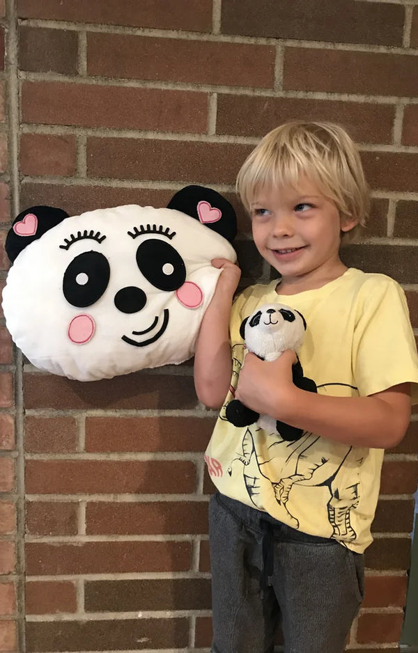A boy stands against a brick wall holding his finished panda pillow and a plush panda