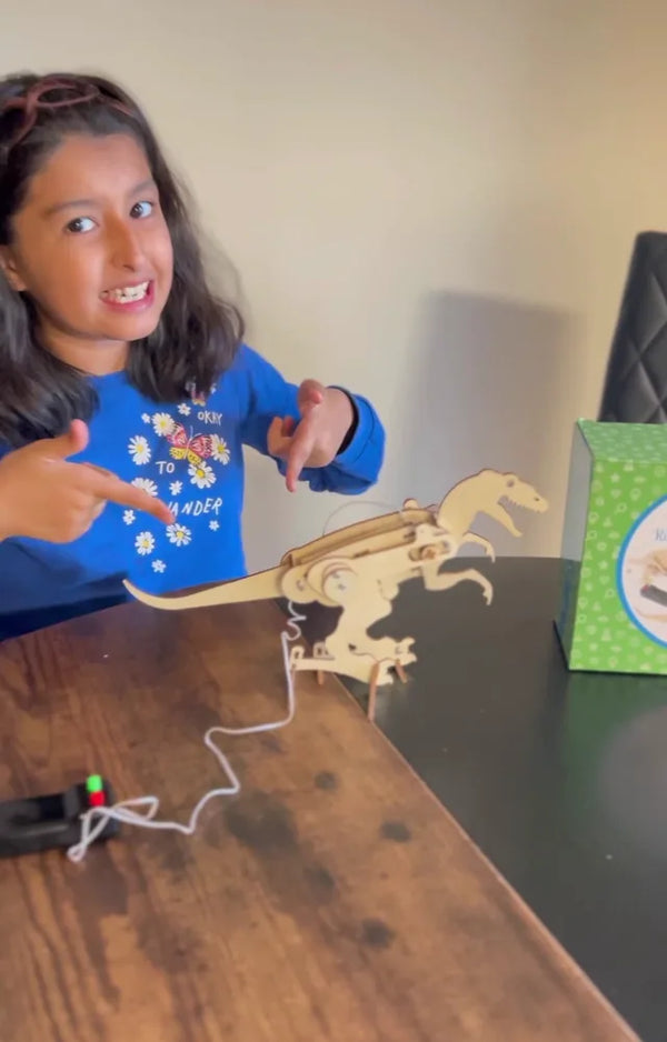 Girl grinning and pointing at her finished wooden T-rex, its controller on the table beside it