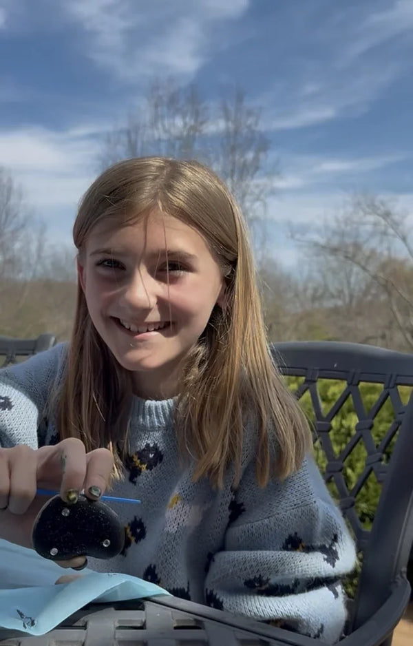 A smiling girl at an outdoor table on a sunny day holding up the dark painted rock she decorated