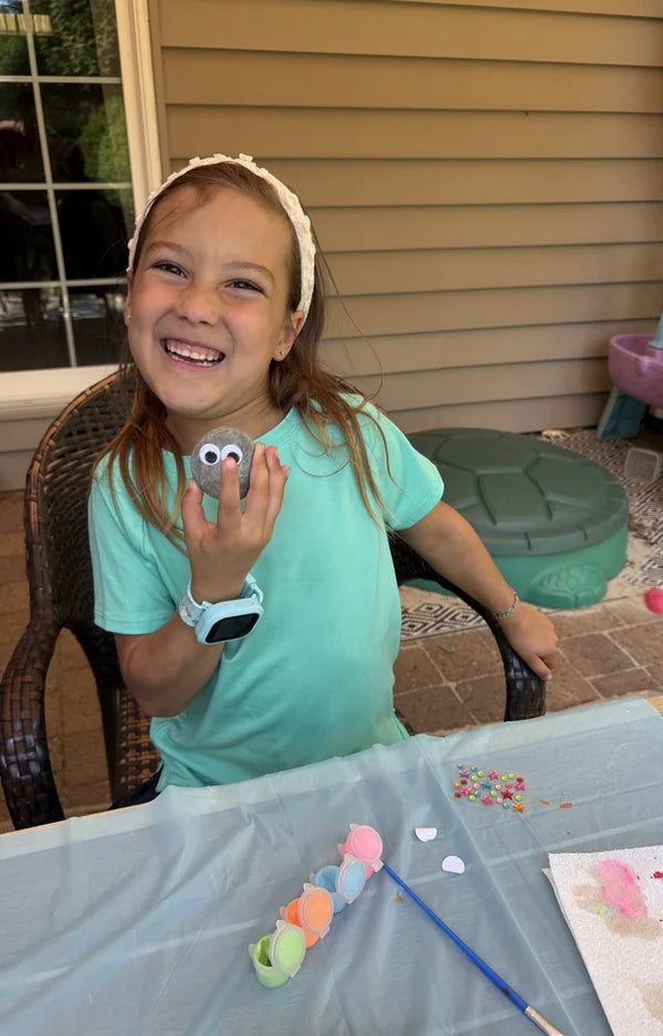 A laughing girl in a teal shirt holding up a googly-eye rock at a painting table on a patio