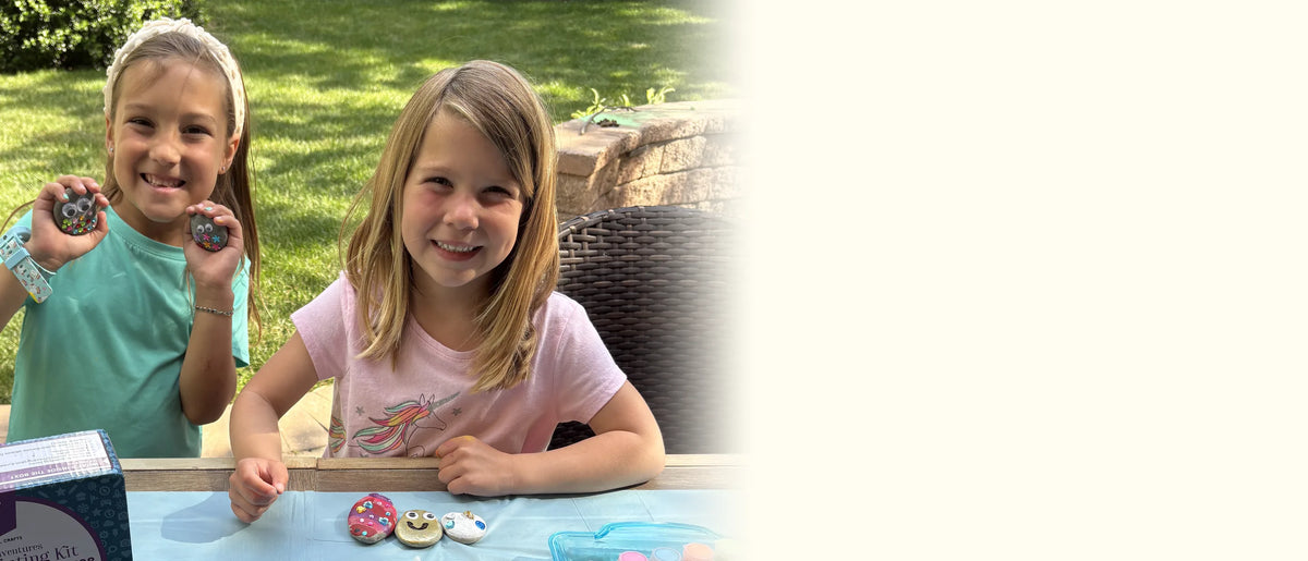 Two smiling girls at an outdoor table holding up painted googly-eye rocks, with the kit box beside them