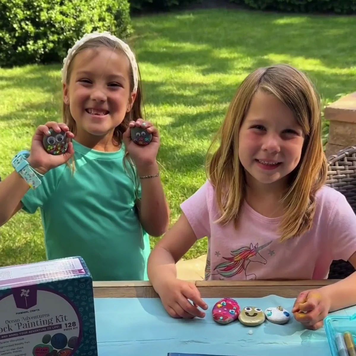 Two smiling girls at an outdoor table holding painted rocks, with the kit box and painted rocks in front