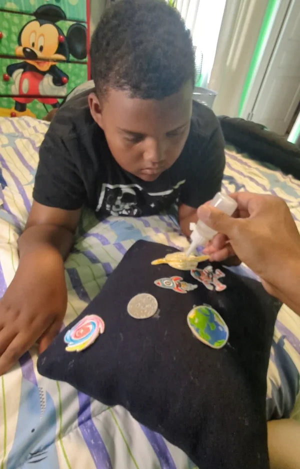 An older boy decorates his space pillow with patches while an adult's hand helps with the glue
