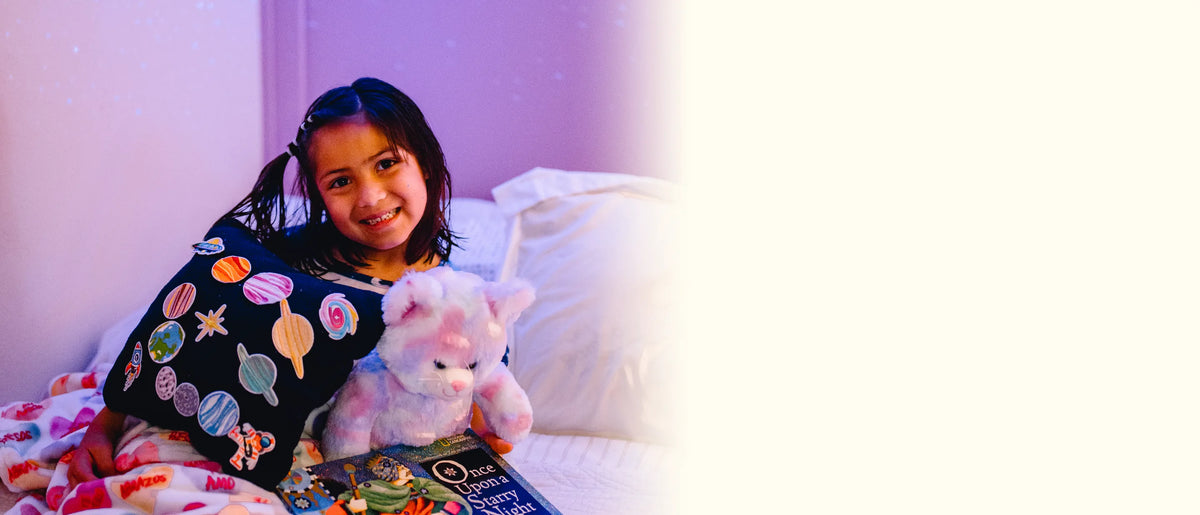 A smiling girl in a purple, star-lit bedroom with her finished space pillow, a plush toy, and a book