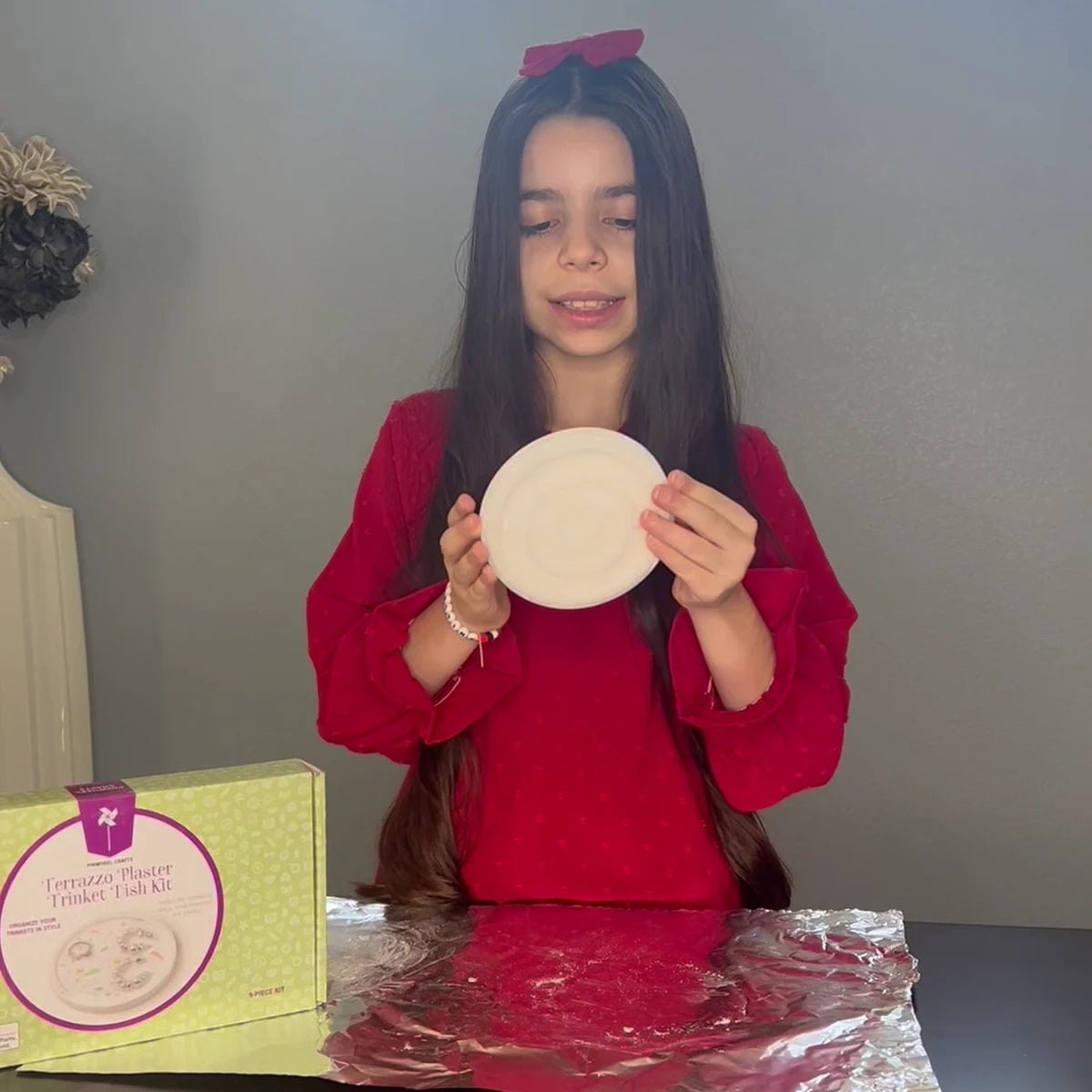 Girl in a red dress holding her finished plaster trinket dish beside the Terrazzo Plaster Trinket Dish Kit box