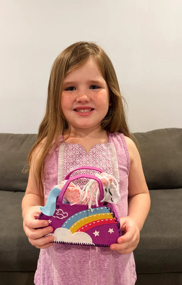 A smiling girl holds up her finished felt rainbow purse with the plush unicorn sitting inside