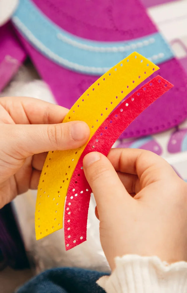 Hands hold the yellow and pink felt rainbow strips, with the pre-punched sewing holes clearly visible