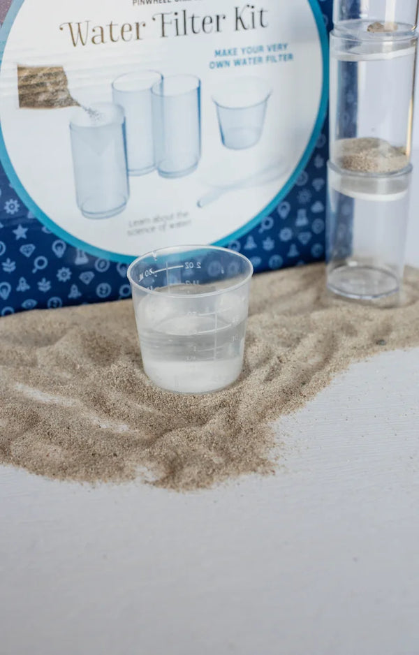 A beaker of water standing on sand in front of the Water Filter Kit box and the filter tower