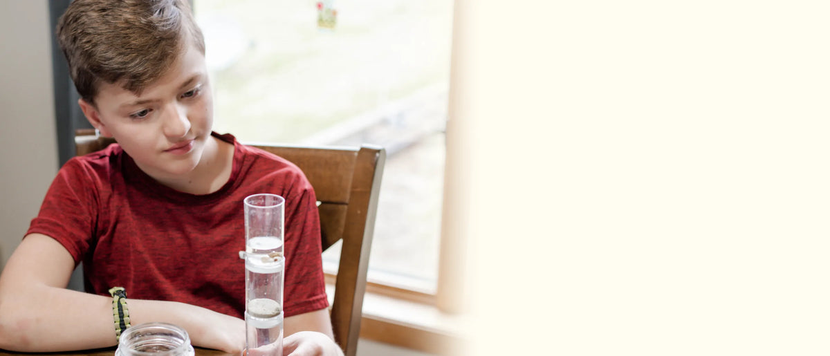 Boy in a red T-shirt at the kitchen table watching his water filter tower, a jar of water beside it