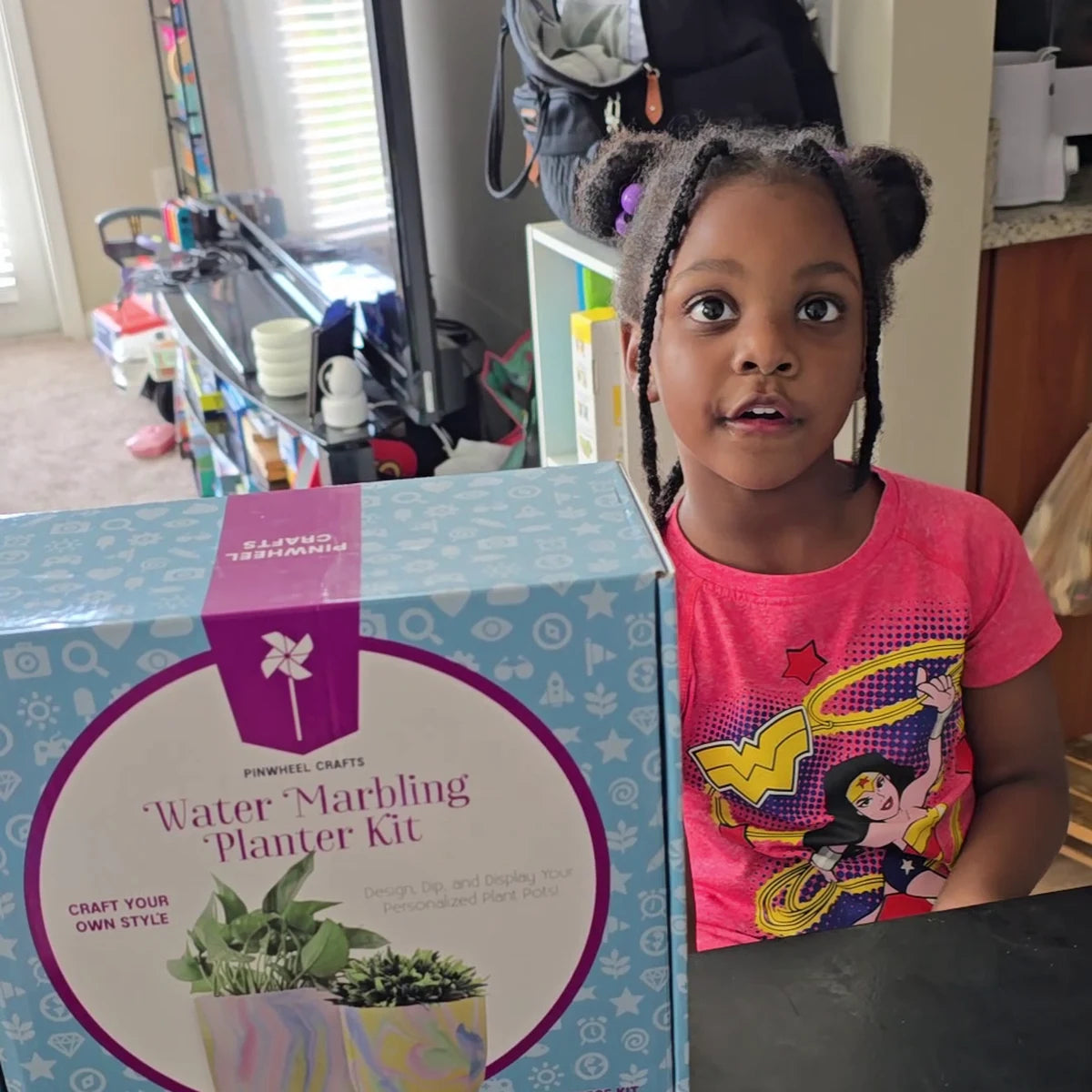 Young girl in a pink T-shirt standing beside the Water Marbling Planter Kit box in her kitchen
